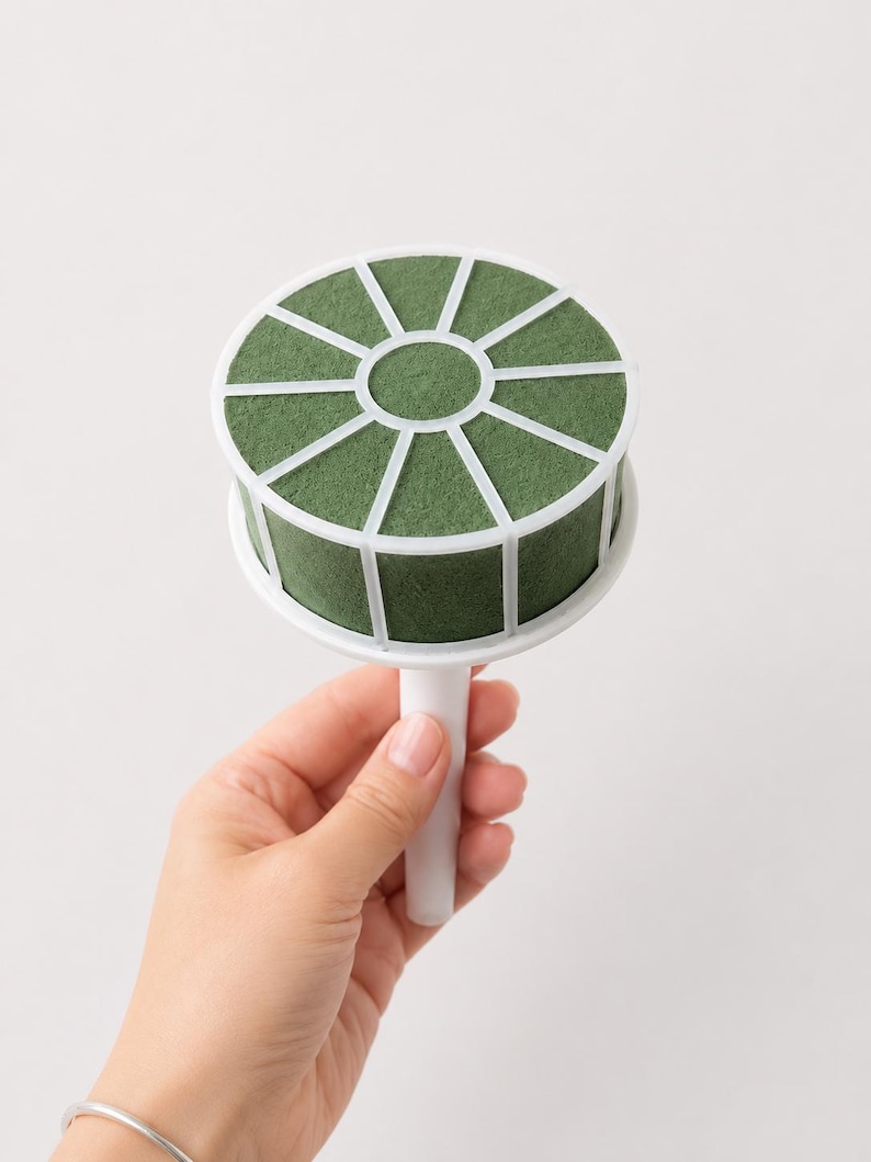 May include: A round floral foam cage with a white plastic frame and handle. The dark green foam is divided into segments by the white plastic grid, designed to hold flower stems securely in place for arrangements and floral displays.