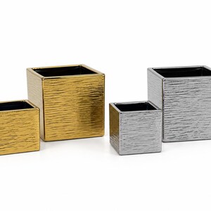 May include: A set of four square planters featuring a textured metallic finish. Two planters are gold and two are silver, with each color pair consisting of one smaller and one larger container. All four planters have a smooth, dark interior.