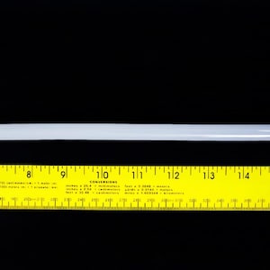 May include: A straight, translucent white cylindrical rod positioned horizontally above a yellow ruler. The ruler displays imperial measurements from 7 to 15 inches, with a conversion chart printed in the center section between 8 and 14 inches.