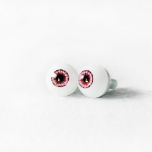 Puede incluir: Dos ojos de mu&ntilde;eca de pl&aacute;stico blanco con iris marrones y detalles morados.