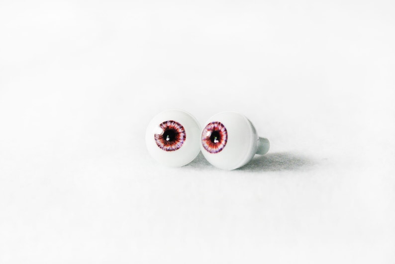 Puede incluir: Dos ojos de mu&ntilde;eca de pl&aacute;stico blanco con iris marrones y detalles morados.