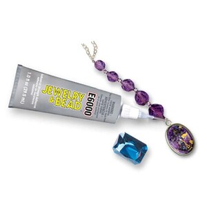 Puede incluir: Un tubo plateado de adhesivo E6000 Jewelry & Bead con una capacidad de 29,5 ml, colocado junto a un collar de cuentas moradas con un colgante ovalado y una cuenta de cristal azul rectangular facetada sobre un fondo blanco.