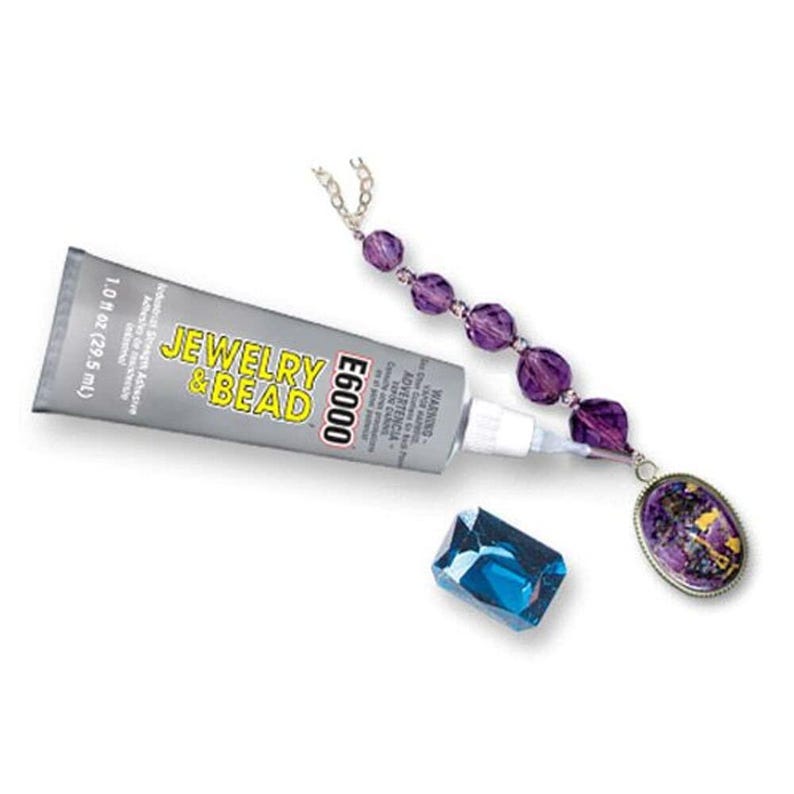 Puede incluir: Un tubo plateado de adhesivo E6000 Jewelry & Bead con una capacidad de 29,5 ml, colocado junto a un collar de cuentas moradas con un colgante ovalado y una cuenta de cristal azul rectangular facetada sobre un fondo blanco.