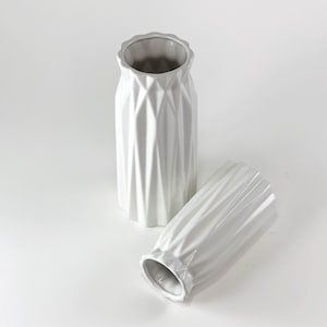 Puede incluir: Dos jarrones de cerámica blanca con un diseño geométrico. Un jarrón está en posición vertical y el otro está tumbado de lado. Los jarrones tienen una superficie texturizada y facetada y un borde festoneado. El interior de los jarrones es de color gris claro.