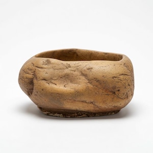 May include: A tan, irregularly shaped bowl with a rough, textured surface that mimics the appearance of a natural stone. The vessel features a wide, open top and a slightly flattened base, showcasing organic contours and subtle variations in color.