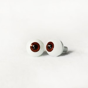 Puede incluir: Dos ojos de mu&ntilde;eca de pl&aacute;stico marr&oacute;n con escler&oacute;tica blanca y pupilas negras. Los ojos est&aacute;n sobre una superficie blanca.