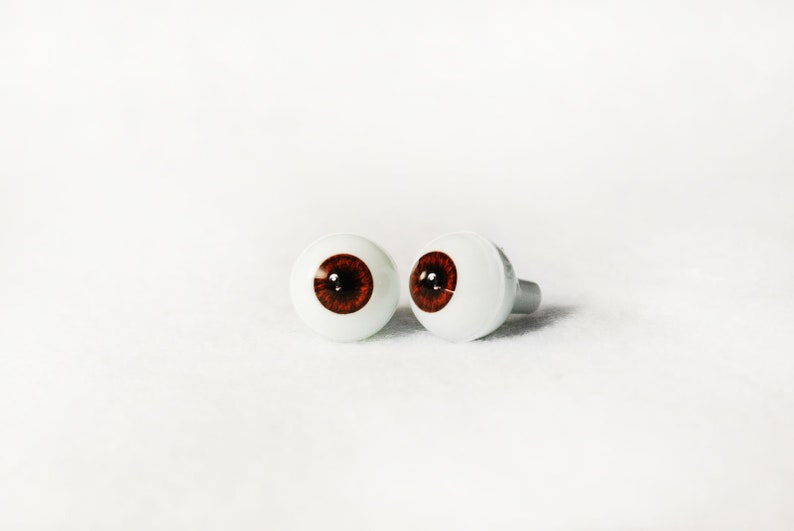 Puede incluir: Dos ojos de mu&ntilde;eca de pl&aacute;stico marr&oacute;n con escler&oacute;tica blanca y pupilas negras. Los ojos est&aacute;n sobre una superficie blanca.