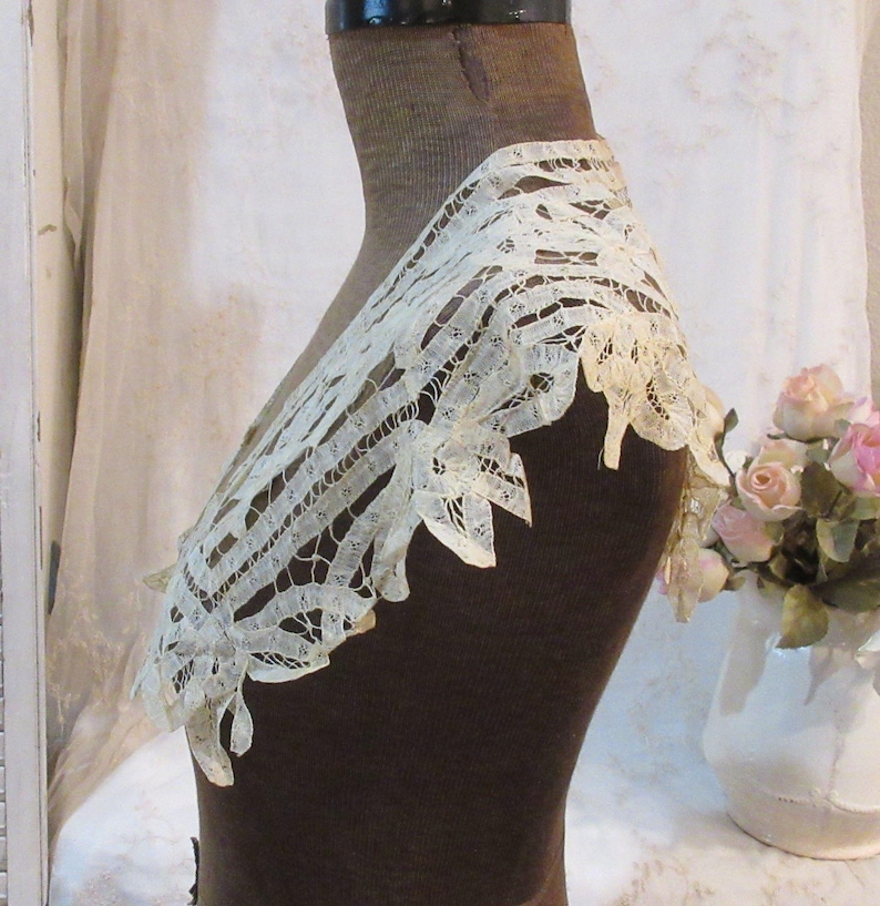 May include: Ivory lace collar with a scalloped edge, displayed on a brown mannequin. The delicate openwork design is visible, showcasing the intricate details of the vintage textile. The collar is a vintage item.