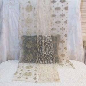 May include: A display of sheer fabric panels with intricate floral and geometric patterns in shades of white, cream, and gold. The panels are draped over a white surface, creating a layered effect. The fabric appears delicate and possibly vintage.