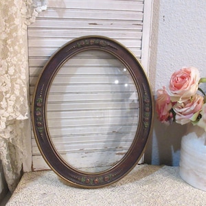 May include: An oval picture frame with a dark brown finish and decorative floral accents. The frame has a gold inner edge and a glass insert. The background includes a white shutter and a vase of pink roses.
