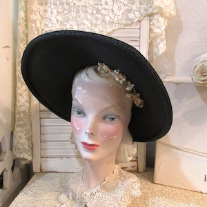 May include: A black hat with a wide brim, adorned with a delicate floral accent, sits atop a mannequin head. The mannequin has painted features, including rosy cheeks and red lips. The hat is a vintage style.