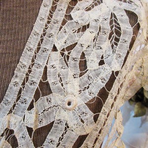 May include: Close-up of cream-colored lace with a floral design. The lace is intricately woven with a delicate pattern, featuring a central flower and ribbon-like details. The background is a textured brown fabric.