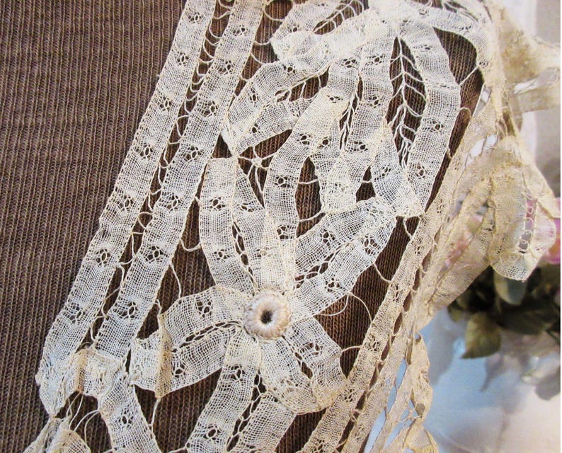May include: Close-up of cream-colored lace with a floral design. The lace is intricately woven with a delicate pattern, featuring a central flower and ribbon-like details. The background is a textured brown fabric.