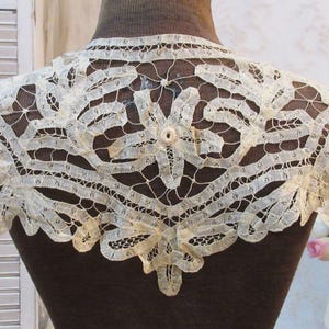 May include: Cream-colored lace collar with a floral and geometric design. The delicate lace is displayed on a brown mannequin, showcasing the intricate openwork and scalloped edges. The collar is a vintage accessory.