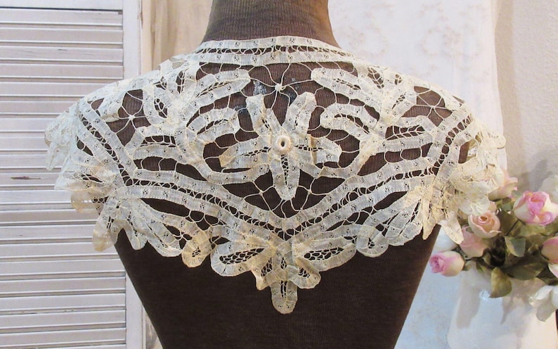 May include: Cream-colored lace collar with a floral and geometric design. The delicate lace is displayed on a brown mannequin, showcasing the intricate openwork and scalloped edges. The collar is a vintage accessory.
