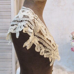 May include: An antique cream-colored lace collar, displayed on a mannequin. The delicate lace features an openwork design with scalloped edges. The collar is likely vintage, showcasing intricate craftsmanship and a timeless aesthetic.