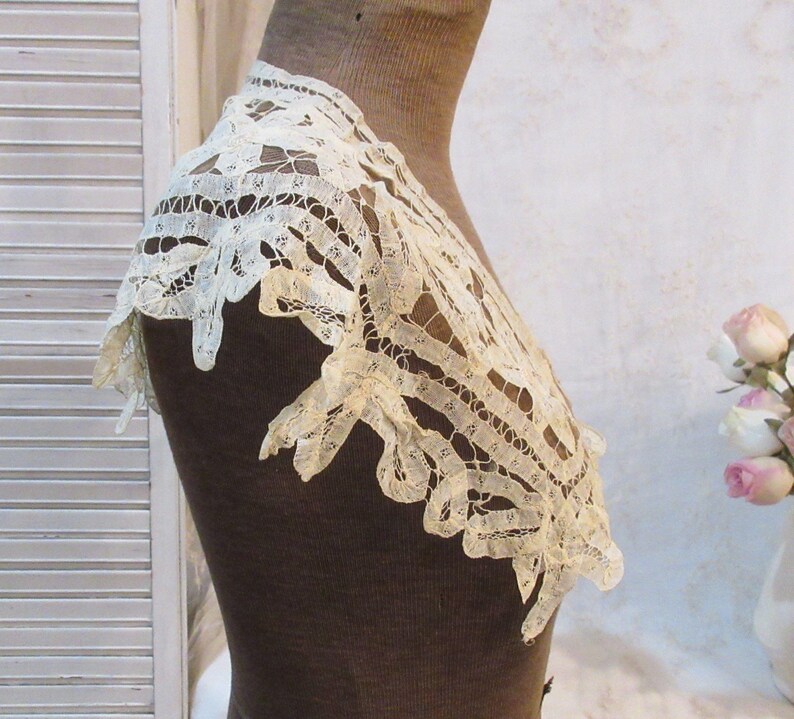 May include: An antique cream-colored lace collar, displayed on a mannequin. The delicate lace features an openwork design with scalloped edges. The collar is likely vintage, showcasing intricate craftsmanship and a timeless aesthetic.