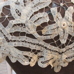 May include: Close-up of an antique, off-white lace collar. The delicate lace features intricate floral and geometric patterns. The collar is displayed on a dark brown surface, highlighting the fine details of the lace.