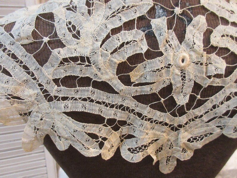 May include: Close-up of an antique, off-white lace collar. The delicate lace features intricate floral and geometric patterns. The collar is displayed on a dark brown surface, highlighting the fine details of the lace.