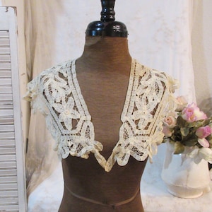 May include: Cream-colored lace collar with floral and leaf patterns. The collar is displayed on a mannequin. The lace has a scalloped edge and a V-neck design. A white pitcher with pink and white flowers is in the background.
