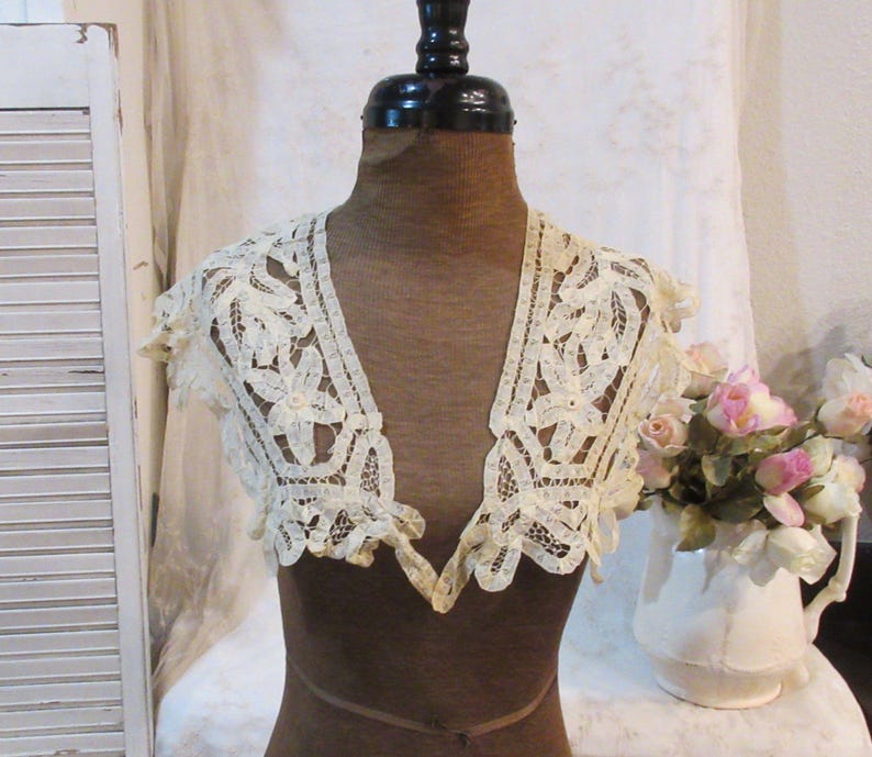 May include: Cream-colored lace collar with floral and leaf patterns. The collar is displayed on a mannequin. The lace has a scalloped edge and a V-neck design. A white pitcher with pink and white flowers is in the background.