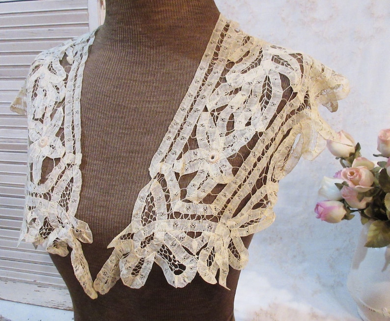 May include: Ivory lace bolero with a delicate openwork design. The bolero features short sleeves and scalloped edges. The intricate lace pattern is visible, showcasing the garment's craftsmanship. This vintage-inspired piece adds a touch of elegance.