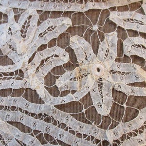 May include: Close-up of antique off-white lace with a floral design. The lace features intricate patterns and a central flower-like motif. The background is a textured brown fabric, providing contrast to the delicate lace.