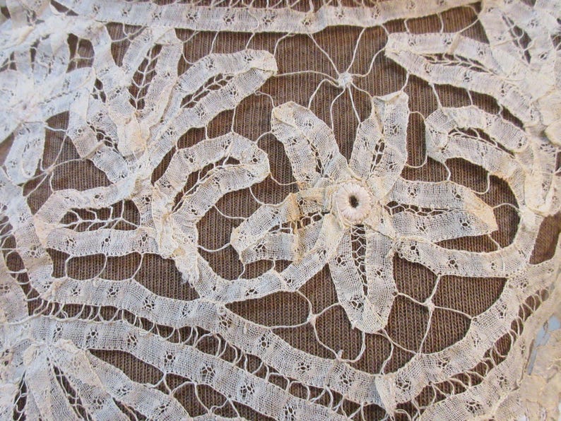 May include: Close-up of antique off-white lace with a floral design. The lace features intricate patterns and a central flower-like motif. The background is a textured brown fabric, providing contrast to the delicate lace.