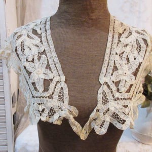 May include: Ivory lace bolero with a delicate floral and geometric pattern. The short-sleeved garment features scalloped edges and a V-neck design. The bolero is displayed on a brown mannequin, showcasing its intricate details and vintage aesthetic.