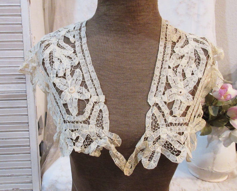 May include: Ivory lace bolero with a delicate floral and geometric pattern. The short-sleeved garment features scalloped edges and a V-neck design. The bolero is displayed on a brown mannequin, showcasing its intricate details and vintage aesthetic.