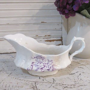 May include: A white ceramic gravy boat featuring a scalloped rim and a delicate purple floral transfer pattern on the side. The vessel has a curved handle and a wide pouring spout, showcasing a glossy finish and an elegant, flowing silhouette.