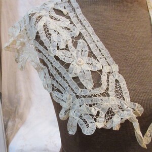 May include: Ivory lace shoulder piece with floral and geometric patterns. The delicate lace features a scalloped edge and a central flower design, creating an elegant and vintage aesthetic. The piece is likely part of a garment.