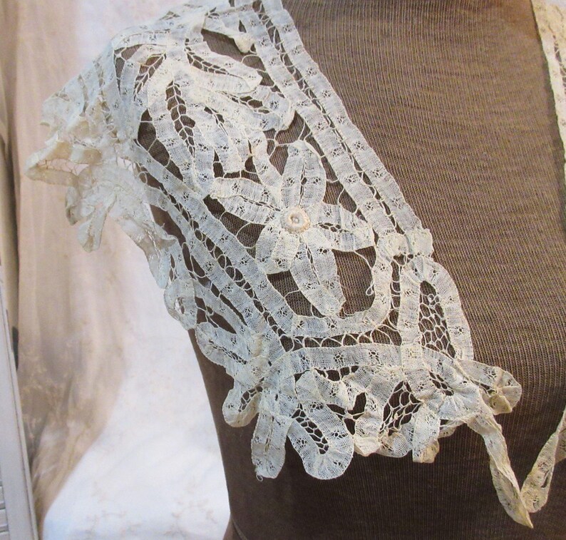 May include: Ivory lace shoulder piece with floral and geometric patterns. The delicate lace features a scalloped edge and a central flower design, creating an elegant and vintage aesthetic. The piece is likely part of a garment.