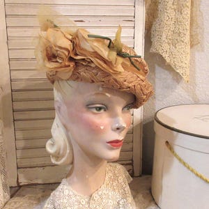 May include: A vintage-style hat with a woven straw base, adorned with large, light tan fabric flowers and a sheer veil. The hat is displayed on a mannequin head with painted features, including red lips and blush. The overall aesthetic is delicate and romantic.
