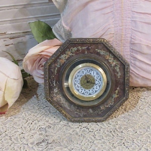 May include: An octagonal clock with a dark brown frame featuring gold-toned floral accents and a central circular face. The clock face has a gold-toned rim, black Arabic numerals, and thin black hands set against a white background.