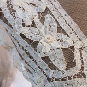 May include: Close-up of a cream-colored lace sleeve with a floral design. The lace is intricately detailed, with a delicate, open weave. A small, round button is at the center of the floral pattern. The sleeve is set against a brown fabric background.