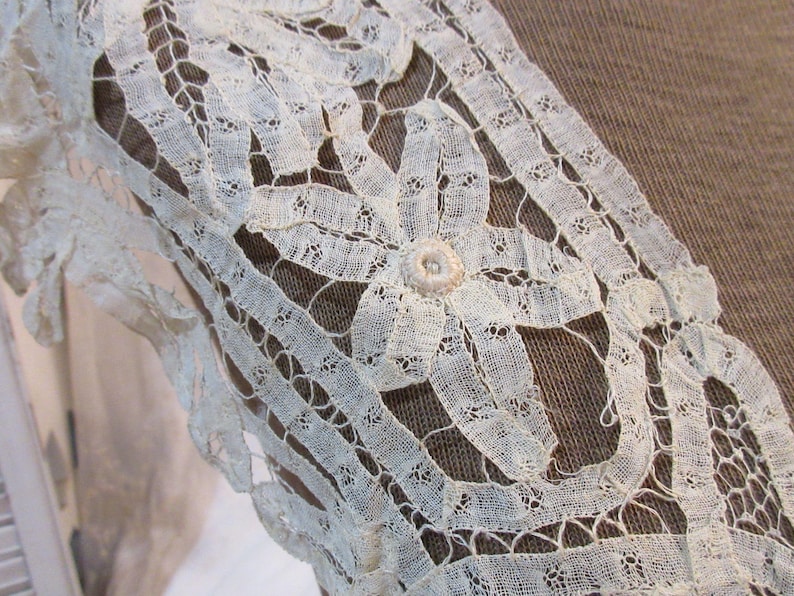 May include: Close-up of a cream-colored lace sleeve with a floral design. The lace is intricately detailed, with a delicate, open weave. A small, round button is at the center of the floral pattern. The sleeve is set against a brown fabric background.