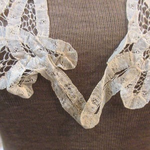 May include: Close-up of an off-white lace collar with a V-neck design. The delicate lace features intricate patterns and a scalloped edge. The collar is set against a brown background, highlighting the lace's texture and detail.