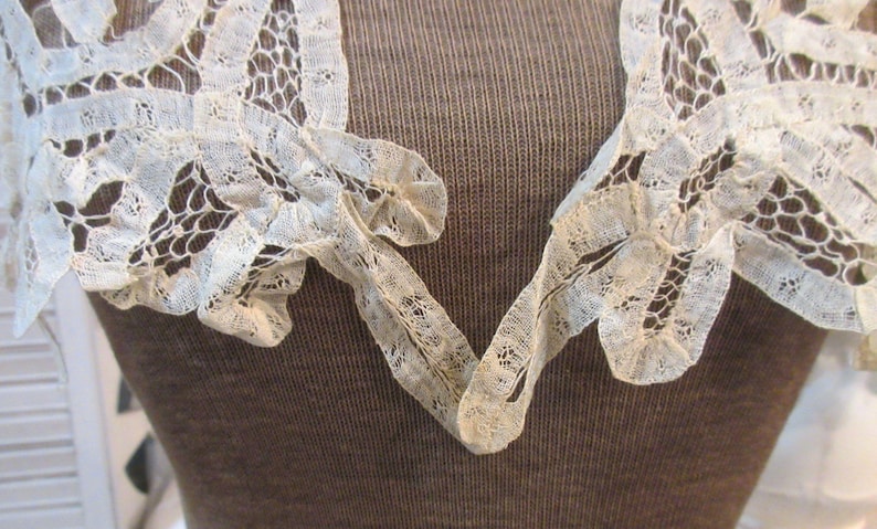 May include: Close-up of an off-white lace collar with a V-neck design. The delicate lace features intricate patterns and a scalloped edge. The collar is set against a brown background, highlighting the lace's texture and detail.