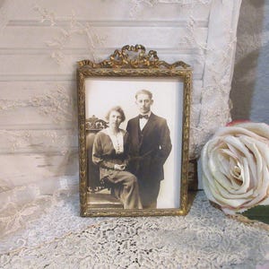 May include: A rectangular gold-toned metal picture frame featuring an ornate ribbon-style crest at the top and a delicate leaf pattern along the border. The frame contains a black and white vintage photograph of two people posing together.