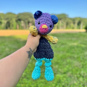 May include: A handmade crochet plush toy in the shape of a bear. The bear has a purple head with blue ears, a pink and yellow beak, and yellow arms. The body is navy blue, and the legs are turquoise. The toy is held against a blurred green background.