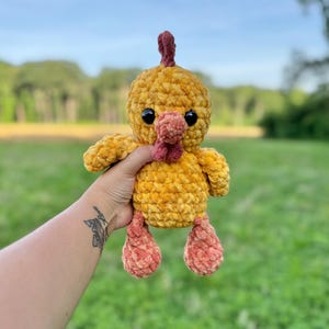 May include: A handmade, plush chicken toy in shades of yellow and orange. The chicken has a textured, fluffy body, black eyes, and a red comb and wattle. The toy is designed to be soft and cuddly, perfect for children.