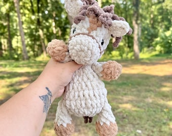 May include: A crochet plush toy shaped like a horse, crafted from soft, chunky yarn in shades of cream, tan, and dark brown. The toy features a textured mane, a defined muzzle, and small ears, with dark stitched eyes and a simple bridle detail.
