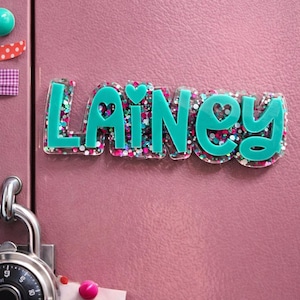 Puede incluir: Una placa con el nombre turquesa que dice "Lainey" en una fuente lúdica, llena de purpurina colorida. La placa con el nombre está fijada a una puerta de taquilla rosa, con un candado de combinación y otros elementos decorativos.