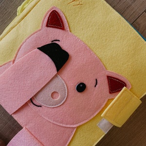 May include: A yellow felt book with a pink felt pig face on the front. The pig has black eyes, a pink nose, and red ears. The book has a flap on the side that says "Happy" in yellow thread.