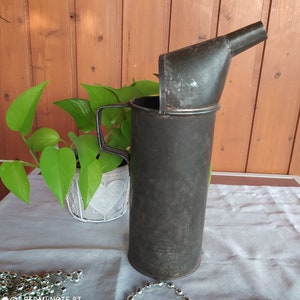 Puede incluir: Un embudo de metal vintage con acabado negro y un pico. El embudo está unido a un recipiente de metal negro con fondo redondeado.