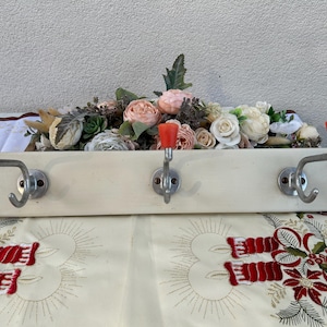May include: White wooden coat rack with three hooks. Each hook has a red plastic knob. The rack is decorated with a floral arrangement of pink, white, and green flowers.