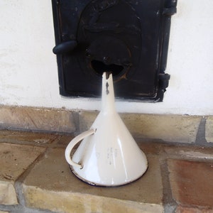 May include: White enamel funnel with a blue rim and a handle. The funnel is sitting on a brick surface in front of a black metal door.