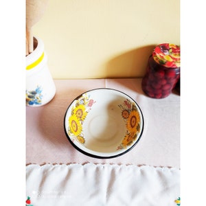 May include: A white enamel bowl with a black rim and a floral design featuring yellow sunflowers, pink flowers, and green leaves.