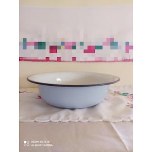 May include: A light blue enamel bowl with a black rim. The bowl is sitting on a white tablecloth with a floral pattern.